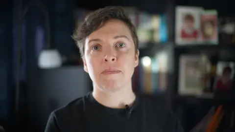 Jamie Niblock/BBC Laura Maisey looks at the camera. You can see her head and shoulders. She has a black t-shirt on and has pale grey eyes. She has short, brown hair swept to one side. There are blurred out pictures of people on the bookshelves behind her. There is some light reflection off the bookshelves.