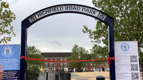 The Highfield Road Park navy blue arch with two Coventry City football club emblems on either side. The red ribbon is attached to either side underneath. Houses can be seen in the background and a park.
