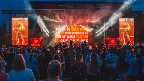 Avril Lavigne performing in Bedford Park in 2024, showing a stage, with screens, her in the middle of the stage, audience members in front, and stage lights. 