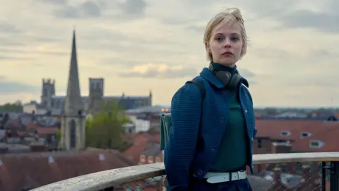 A woman with headphones around her neck stands on a balcony overlooking York city centre and York Minster.