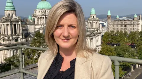 Grainia Long with Belfast City Hall behind her. She is wearing a beige jacket and black top. She has blonde hair.