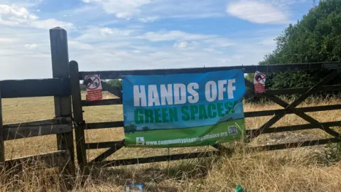 Photograph of a gate with a protest sign against the housing development attached. The image shows a wooden gates, with farmland and trees in the background.