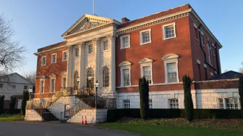 A view of Warrington Town Hall. It is a Georgian building with a large staircase leading up to the front. The centre of the building at the front has four white pillars.
