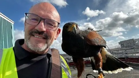 Andy Crowle smiling for the camera with one of his Harris hawks, Willow. They appear to be on the rood of a tall building. It is a cloudy day.