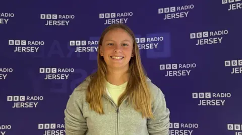 Lauren wearing a grey hooded jacket. She is smiling at the camera and is stood in front of a purple Radio Jersey sign.