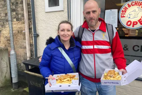 BBC News A woman wearing a blue coat and a man wearing a red and grey tracksuit top stand outside a fish and chip shop, both holding a portion of golden fish and chips in a white cardboard box. The woman has blond hair, tied back, while the man has a shaved head and salt-and-pepper beard. The chip shop has a round white sign in the window showing a smiling blue cartoon fish and the red words: "Fish and Chips, Open".