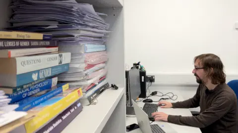 A man with long brown hair and a beard sitting at his desk working on a computer. He is wearing a brown top. To his left is a bookcase with a selection of psychology books. The walls are white.