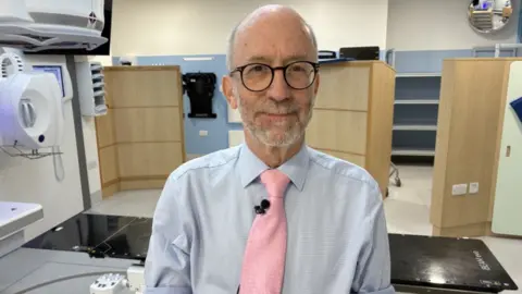 A man pictured in a clinical setting. He has balding hair, is wearing glasses and a light blue shirt with a pink tie. 