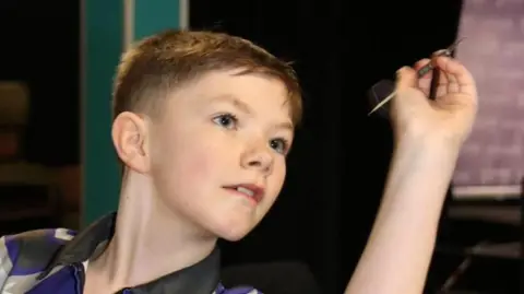 George, who has short brown hair, looking focused as he throws a dart with his left hand.