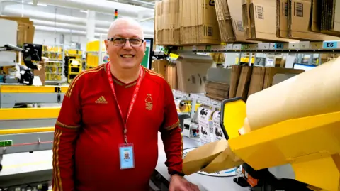 Inside a large warehouse packing area, a Darren Wells is wearing a red sports top with an Adidas logo and a lanyard. He stands near a workstation. The foreground shows a yellow packing machine dispensing brown paper, while shelves above are stacked with flat cardboard boxes labeled with codes like “E4” and “E6.” The background reveals more shelving, conveyor belts, and industrial lighting.
