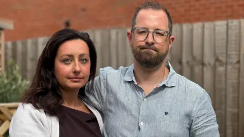 A woman and a man stand in front of a wooden fence, pictured from the shoulders up. The woman has shoulder-length dark hair and wearing a brown top and grey cardigan, and the man is wearing a blue shirt and glasses.