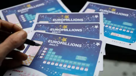A hand holding a pen above five blue EuroMillions tickets. 