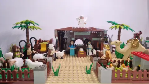 Northlew Nativity Exhibition A nativity scene made of lego. There are lego mini figures of Mary and Joseph and the Shepard's and Kings. There are lego animas including sheep, chickens and a camel. The background is  grey wall. 