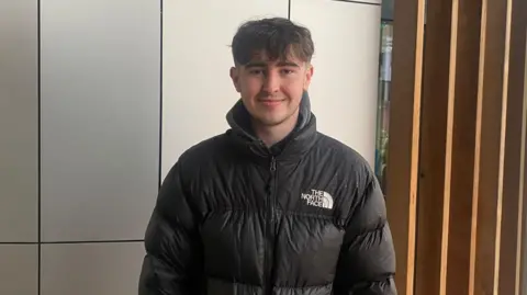 Handout Matthew Lounton wears a black North Face puffa jacket. He has brown hair and is standing in front of a wall with silver panels.  