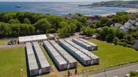 Duke Marketing An aerial view of rows of portable cabins in a field, there is a large marquee in the background, you can see Douglas bay behind, on a clear day.