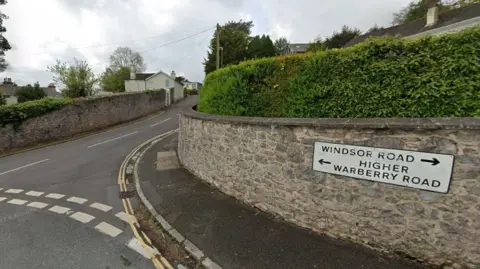The end of a road with a sign fixed to a brick wall that points to Higher Warberry Road in Torquay. 