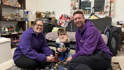 Two Community care nurses from the charity Jessie May, wearing purple jumpers. They are sitting in a Christmassy living room with Alex, a boy they help. Alex has a cystic hygroma, a facial growth that can affect his breathing and eating. They are playing with Doctor Who toys.