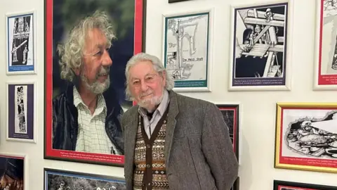 MANX NATIONAL HERITAGE Shane Lucas, a man wearing a tweed jacket, he smiles and stands in front of a portrait of himself and various black and white sketches of the mine.