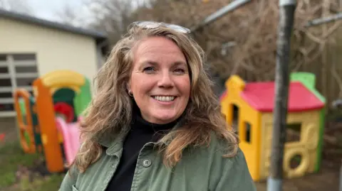 Steve Hubbard/BBC Suzi has fair and curly hair falling below shoulder length. She has a big wide smile into camera and is wearing her glasses on her head and silver-hooped earrings. She's outside in a playground setting with children's toys in the background.  Suzi is wearing a black woollen sweater and an olive green jacket.    