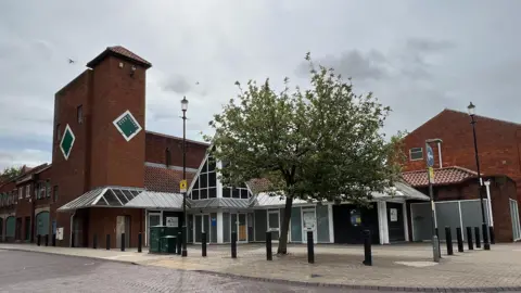 BBC The City Square shopping centre, which has been closed since 2019. It is a red-brick building with a three-storey tower and angular window features. Outside is a tiled square, dominated by a tree with green leaves.