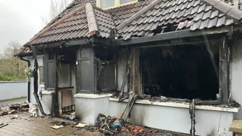 A view of the house which has been gutted in the fire - it is a chalet-bungalow style and the front door and front window are completely gone, surrounded by charred frames. Inside the living room it is black with soot. Guttering has collapsed and debris lies on the paved driveway. It is raining.