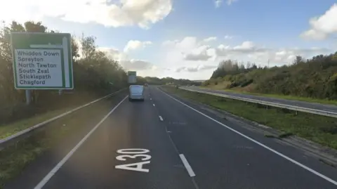 Vehicles drive along the A30 near the junction for Whiddon Down, Spreyton, North Tawton, South Zeal, Sticklepath and Chagford on a sunny day with white clouds in the sky.