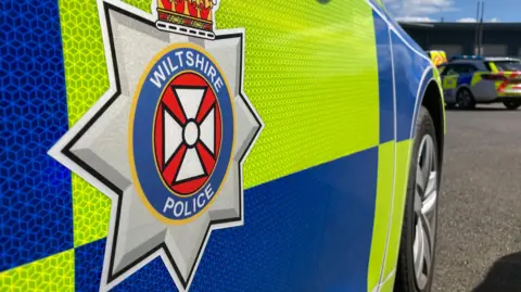 BBC The side of a Wiltshire Police car. Police cars are also parked up in the background of the image.