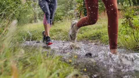 Two sets of women's legs running on a muddy path with green grass at either side. 