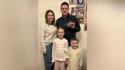 Family photo A family image of mum, dad and the two daughters. Mum Naomi is in a beige jumper and jeans, dad Wayne is in a black, zip up top, the older daughter Imogen is in a pink, yellow and blue striped jumper and Delilah is in a yellow/gold dress and cardigan.