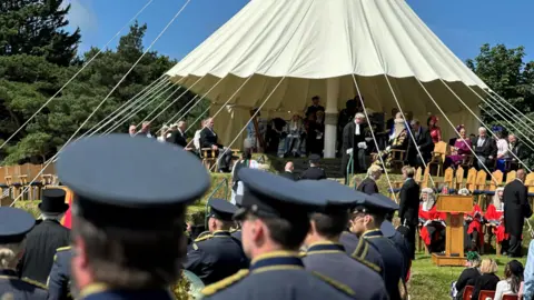 The military band in front of Tynwald Hill, which is surrounded by wooden chairs and has a white canopy over the top.
