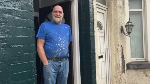 Jonny Humphries/BBC Ken, who is bald and has a grey beard, and wears a blue t-shirt and jeans spattered with white paint from his work, stands in the doorway of a terraced house with the brickwork painted dark green and smiles at the camera. 