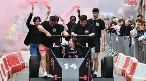 Hinckley BID A group of people, some with flares in the background, on a race track pushing a black soapbox car with pink flashes