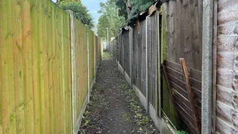 BBC/ Alex James A photo of an alleyway with wooden fences either side. The ground is uneven and muddy. On the right one of the fences has been kicked in and has collapsed 