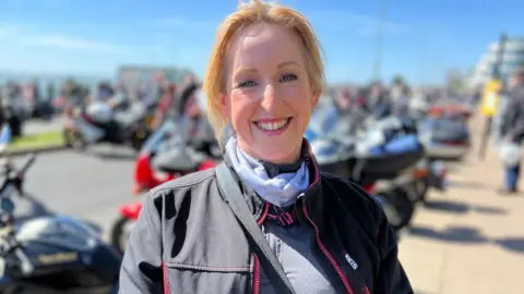 Stuart Woodward/BBC Lorraine Hudson wears a black jacket over a grey top and light blue scarf, as well as a bag strap over her shoulder and chest. She has blonde hair and is smiling at the camera. There are motorbikes blurred out behind her.