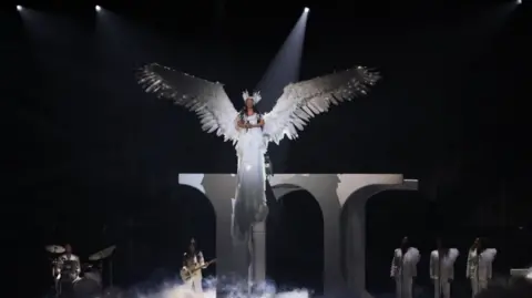 EPA Jade Thirlwall performs during the 2025 BRIT Awards ceremony at The O2 arena in London. She wears huge white wings and a winged headband. She is dressed all in white and is lifted above white columns while a band also dressed in white plays below.