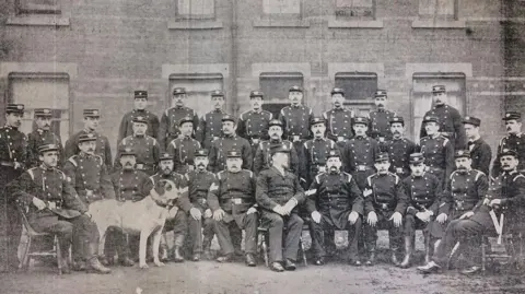 A formal black and white group photograph showing a large uniformed brigade posed outdoors in front of a brick building. The group is arranged in several rows, with some people seated on wooden chairs at the front and others standing behind them.
