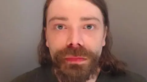 Mugshot of Benjamin Clarke. He has a thick dark goatee beard and long dark hair, and is wearing a black t-shirt.