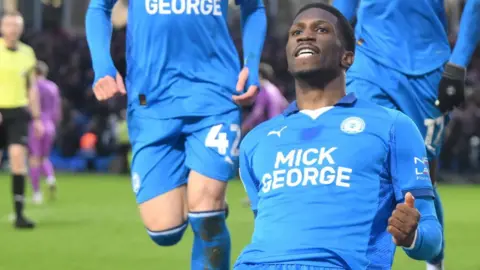Getty Images Pemi Aderoju kneels on the ground in celebration after scoring a goal. He is wearing a blue football kit with "Mick George" branding. 