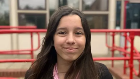 Elliot Deady/BBC A schoolgirl - Amelia - smiling straight at the camera while standing in front of a school building. She is wearing her school uniform - a black blazer over a red and white checked blouse.