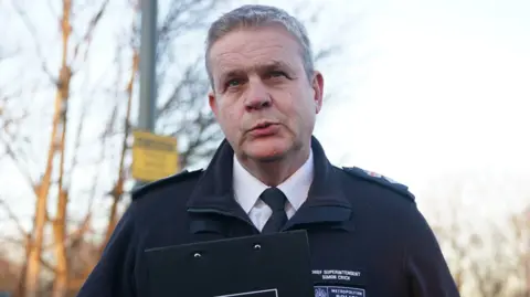 A male, uniformed police officer with short grey hair, mid-speech, holding a black clipboard. His name tag, on his uniform, reads Chief Superintendent Simon Crick. 