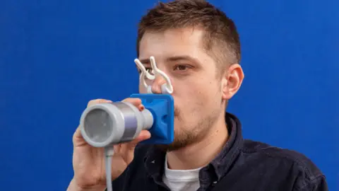 Children of the 90s A young man blowing into a hand-held medical device, standing against a plain blue background