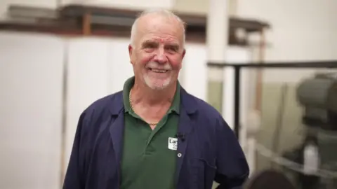 Colin Stevens has short white hair, with a white goatee beard. He wears a dark blue boilersuit over a green polo shirt. He has a gold chain round his neck.