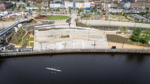 Stockton Council An aerial view of the white steps of the amphitheatre which faces on to the river. There are two people rowing a boat in the water. there are winding paths and greenery dotted around the development.