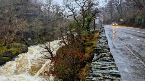 BBC Weather Watchers A flooded road to the right, and on the left a fast-flowing river 