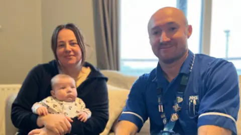 12-week old Myles is wearing a white baby grow and is being held by his mother. His mother, Shanice, is wearing a black zip hoodie and has brunette hair. Sat next to her is Chris in blue nurses scubs. He is smiling and has very short hair.