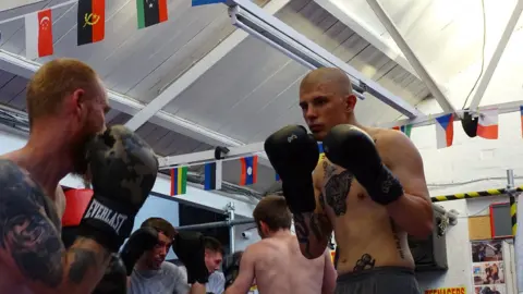 Daniel Bainger Daniel taking part in a boxing exercise, with four other people boxing. He has no top on, is wearing shorts and has tattoos on his body. The room has flags behind him.