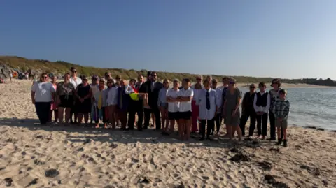 The sea swimming group stand on a sandy beach. The swimmers are standing in a row wearing office wear.
