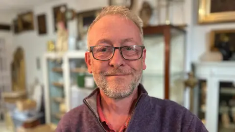 George Carden/BBC Henry standing in his shop with cabinets and paintings behind him. he has grey hair glasses and a three quarter zip 