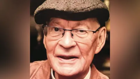 Close up photo of Right Reverend Stephen Pedley. He's wearing a brown flat cap, glasses a brown jacket and red jumper with a clerical dog collar around his neck.