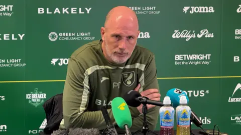 Steve Hubbard/BBC Philippe Clement sits at a table with microphones in front of him during a press conference at Norwich City. He is bald with some stubble on his chin. He wears a green Norwich City branded top and has his arms crossed in front of him.